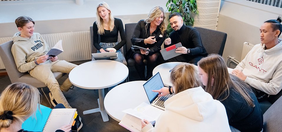 Elever sitter och pluggar i fåtöljer. En lärare samtalar med en elev.